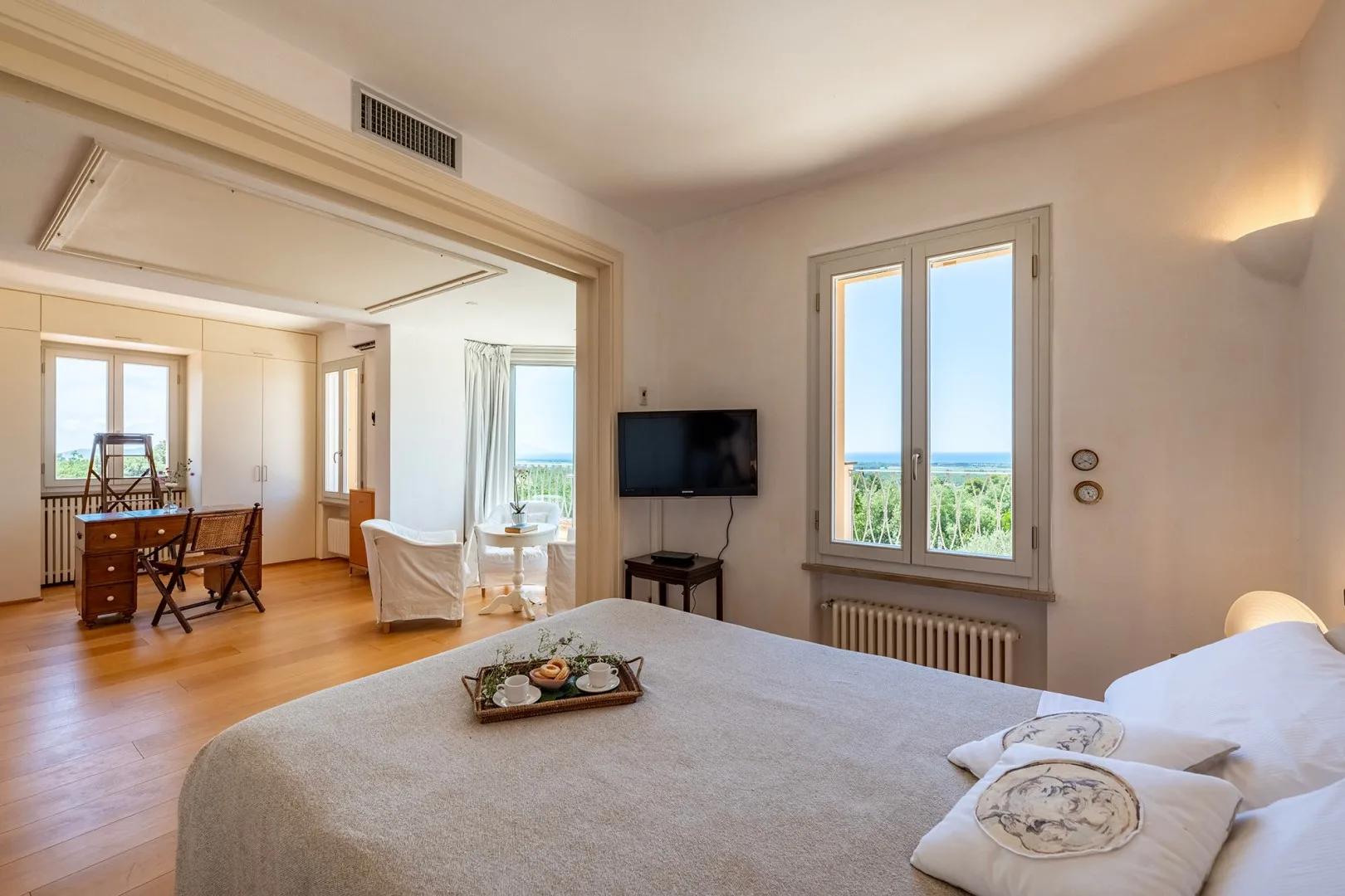 Un dormitorio con cama, televisor y ventanas altas con vistas a vegetación y agua.