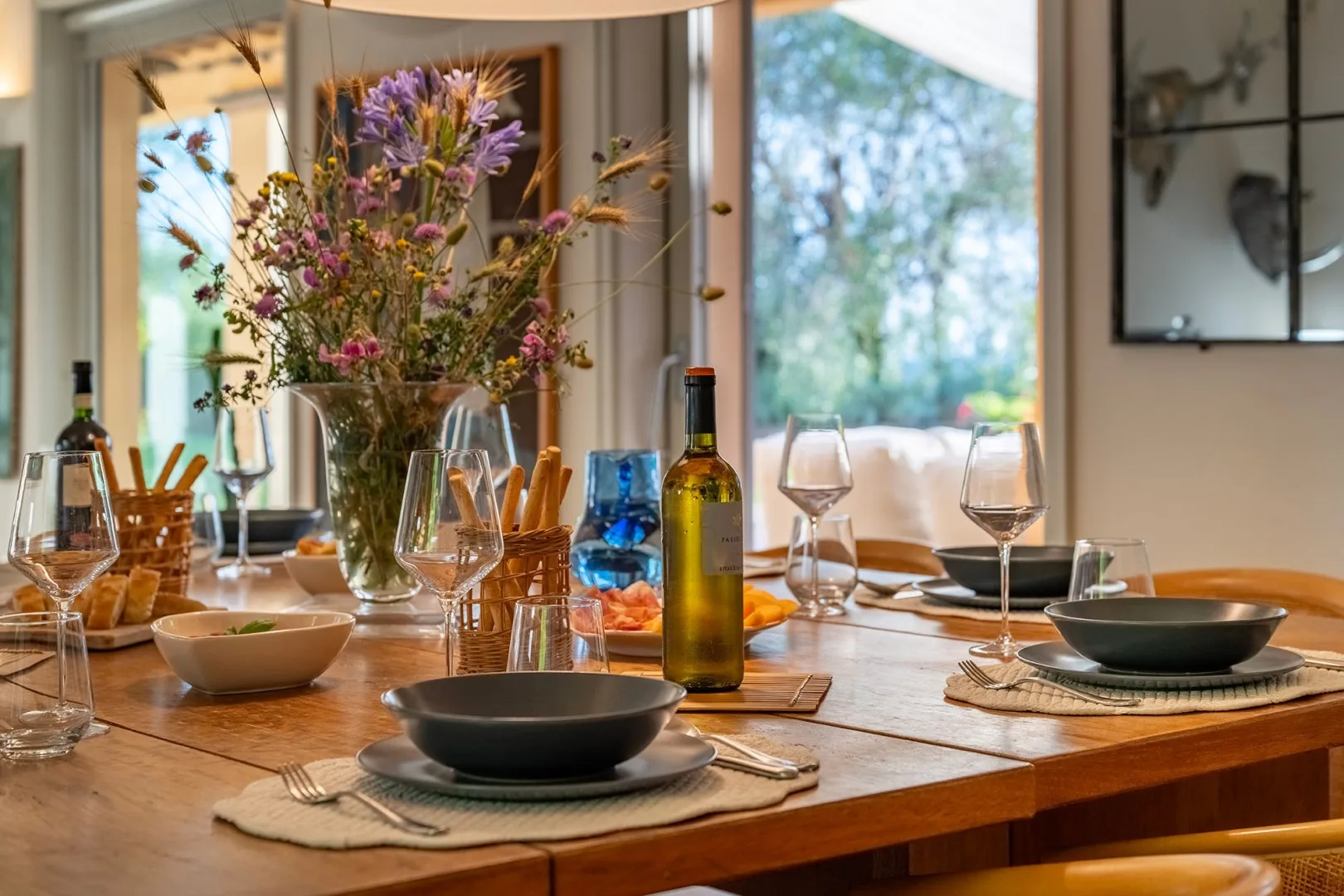 Una mesa de comedor está preparada con platos y copas de vino junto a un jarrón con flores.