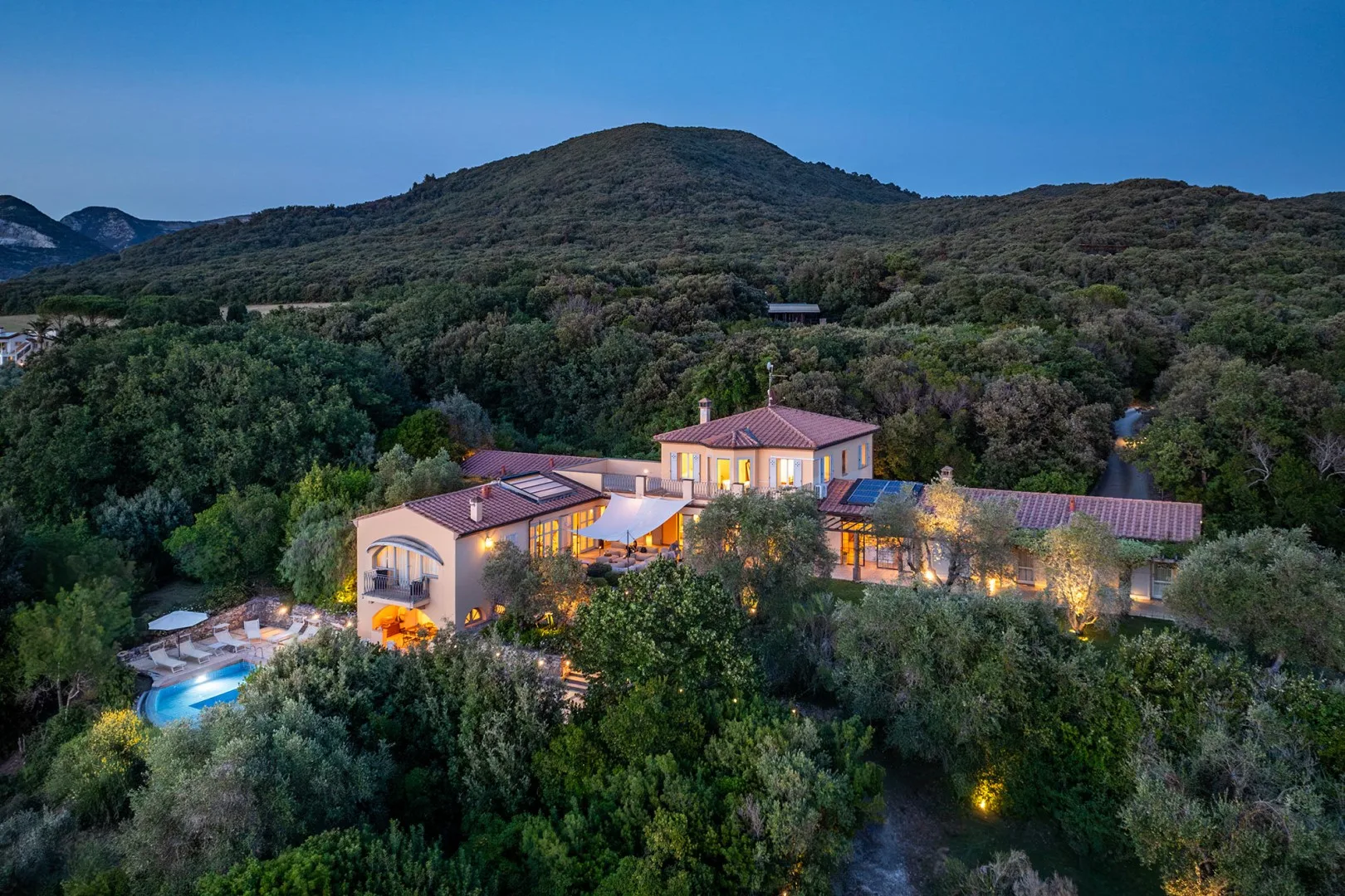 Una casa grande está entre colinas boscosas, con una piscina a su lado.