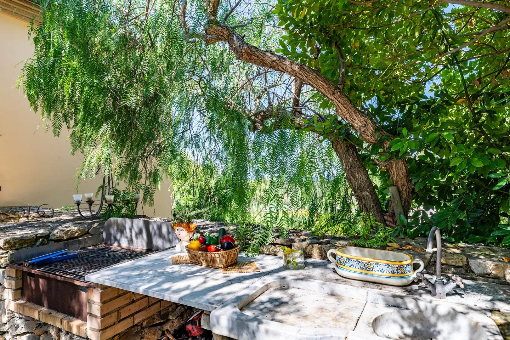 Una encimera de piedra al aire libre está bajo árboles frondosos, junto a una cesta de fruta.