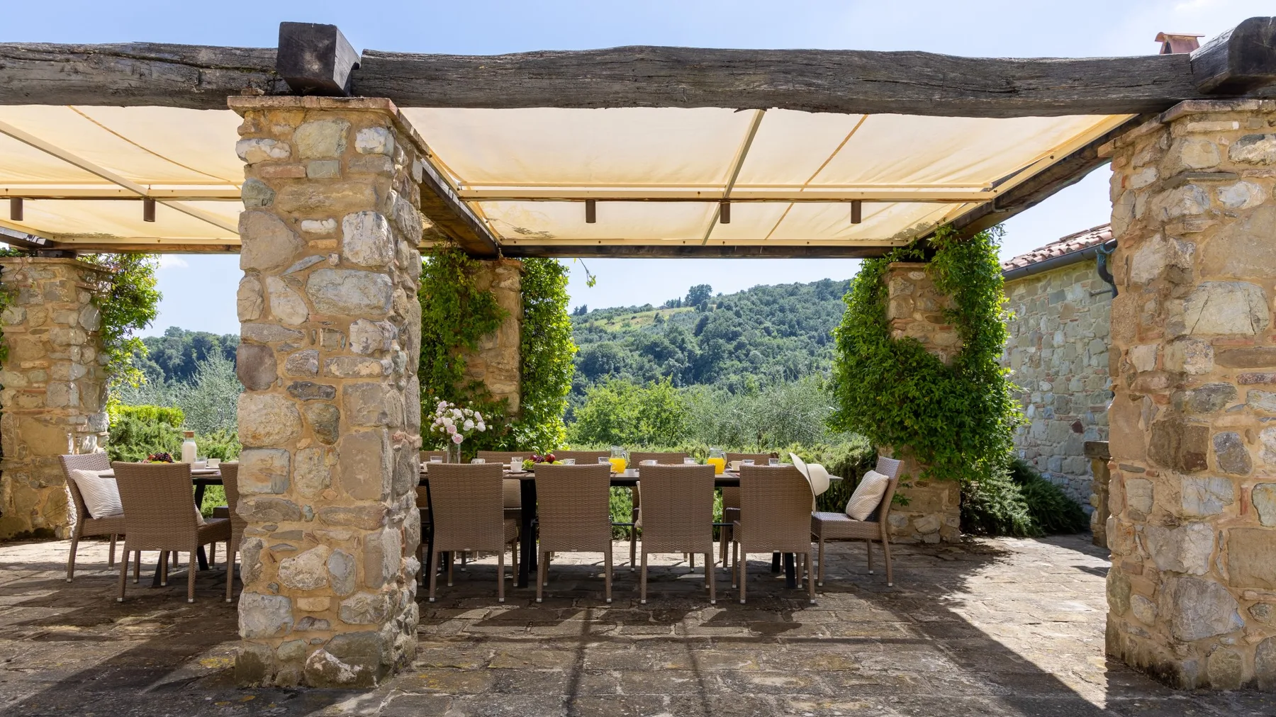 Una zona de comedor al aire libre con mesas y asientos bajo una terraza de piedra sombreada.