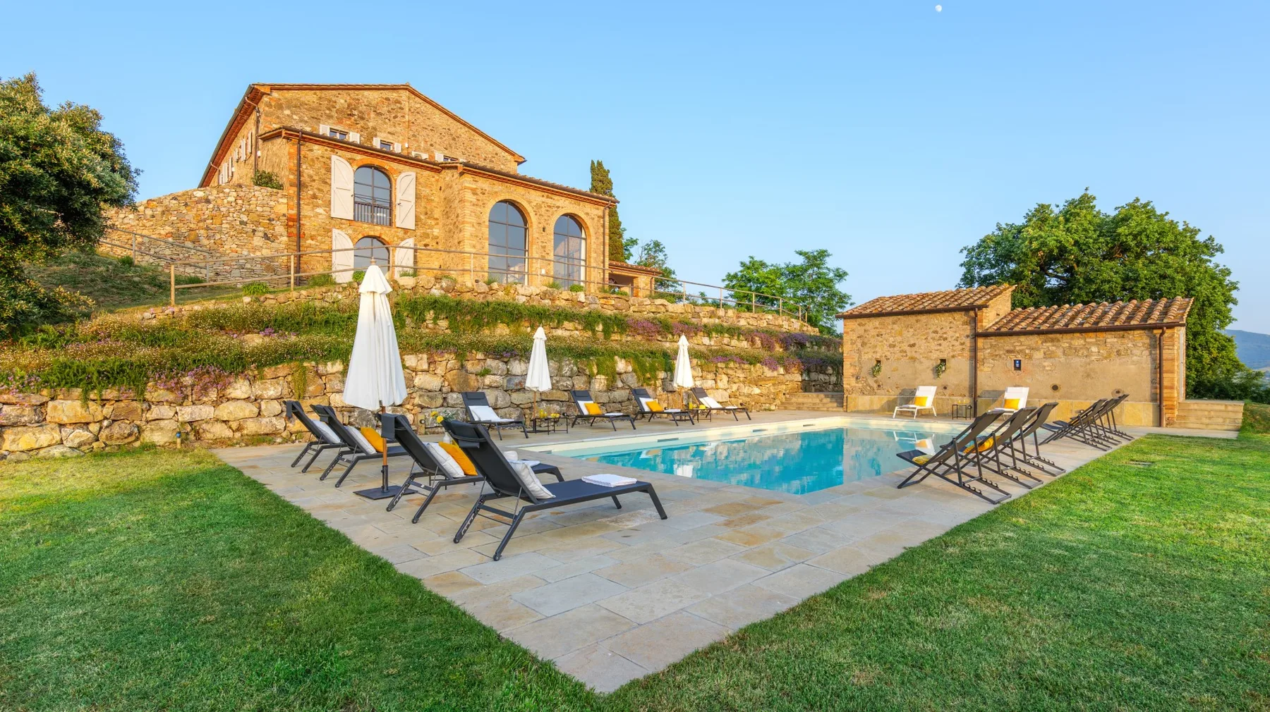 A swimming pool and lounge seating sit beside a stone building and grassy lawn.