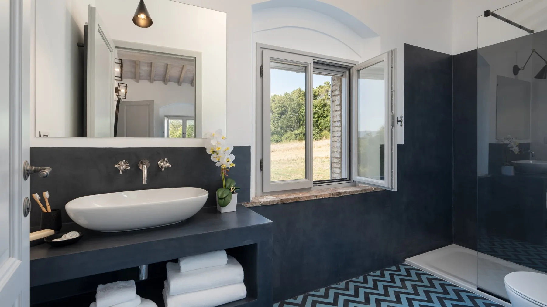 A bathroom with a dark vanity, basin, and open window overlooking greenery.