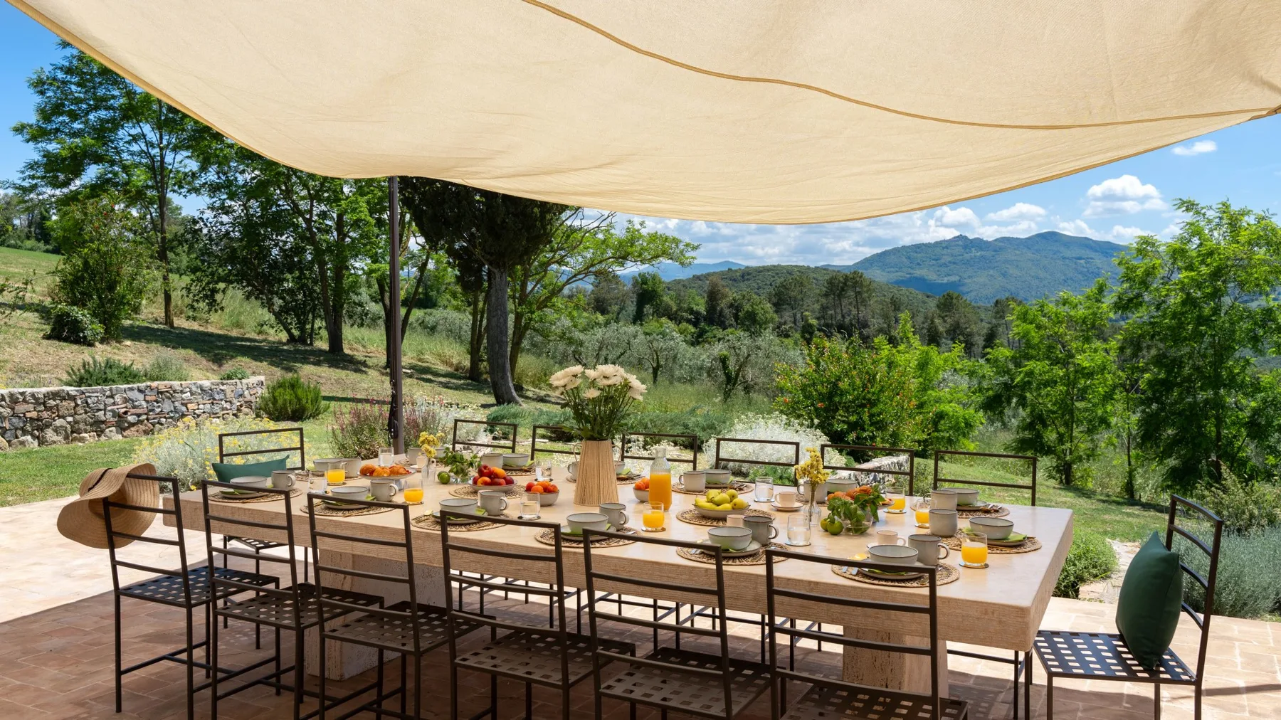 An outdoor dining table is set beneath a shade canopy, with green hills in the background.