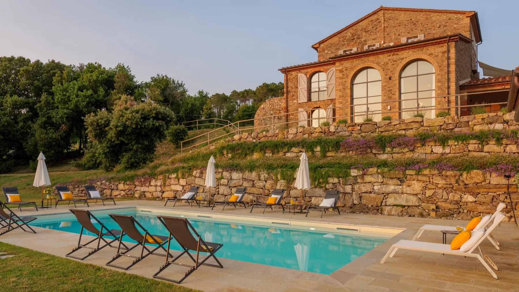 An outdoor swimming pool with lounge seating sits below a brick building and landscaped terrace.