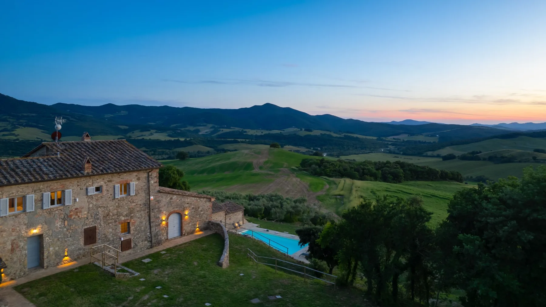 A stone building sits beside a swimming pool amid rolling green hills at dusk.