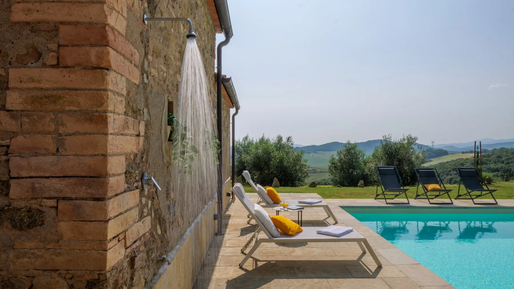 A swimming pool beside a stone patio with lounge chairs and a wall-mounted shower.