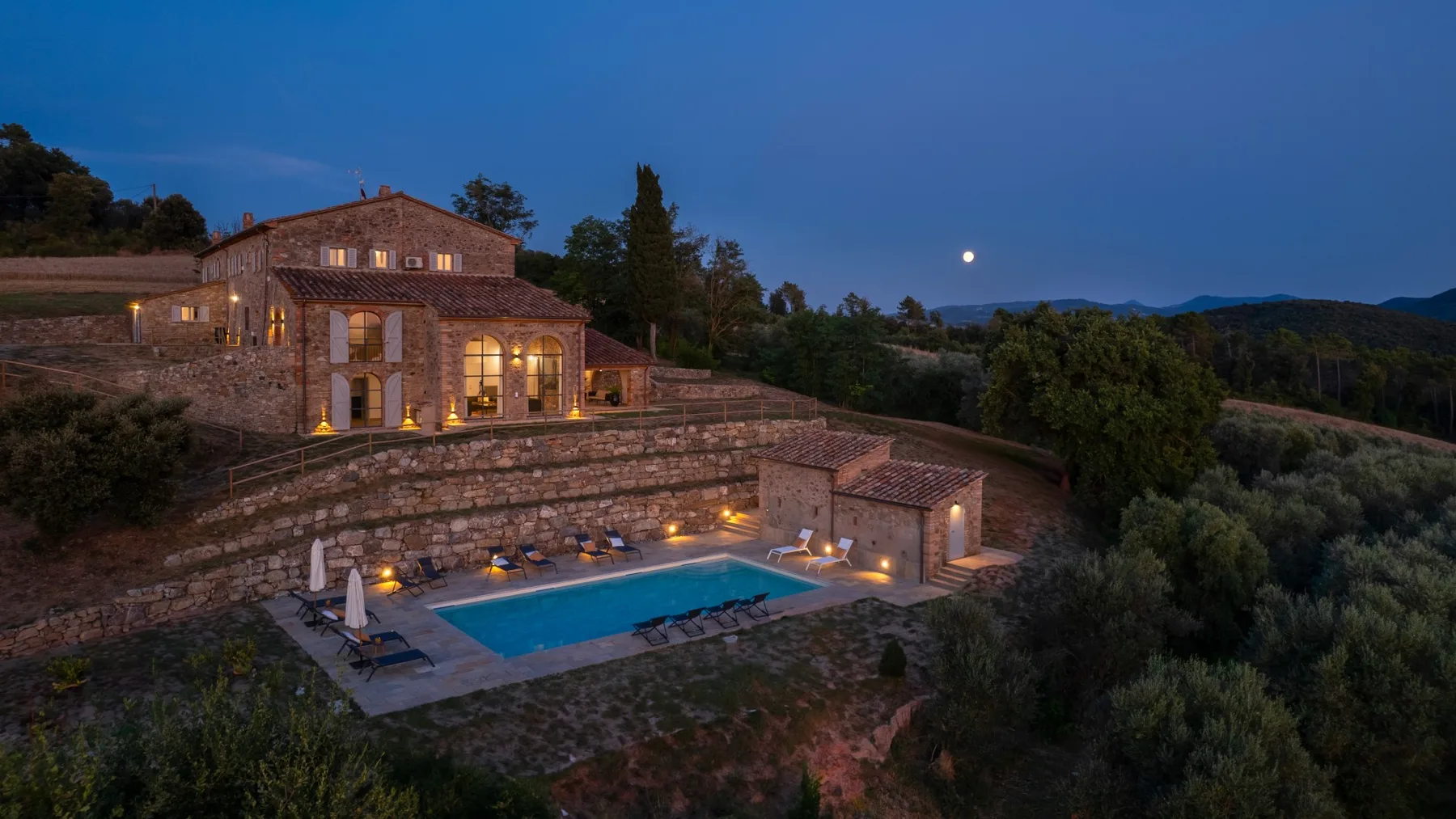 A stone country house overlooks a lit swimming pool at dusk.