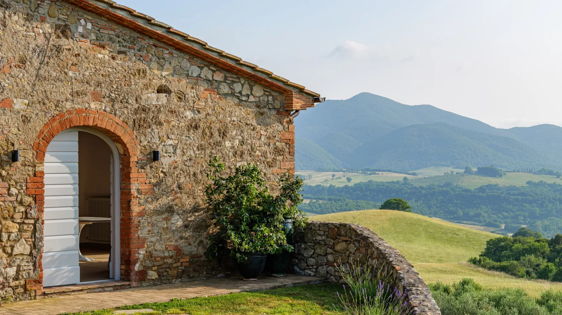 A stone building opens onto a terrace overlooking green hills and distant mountains.