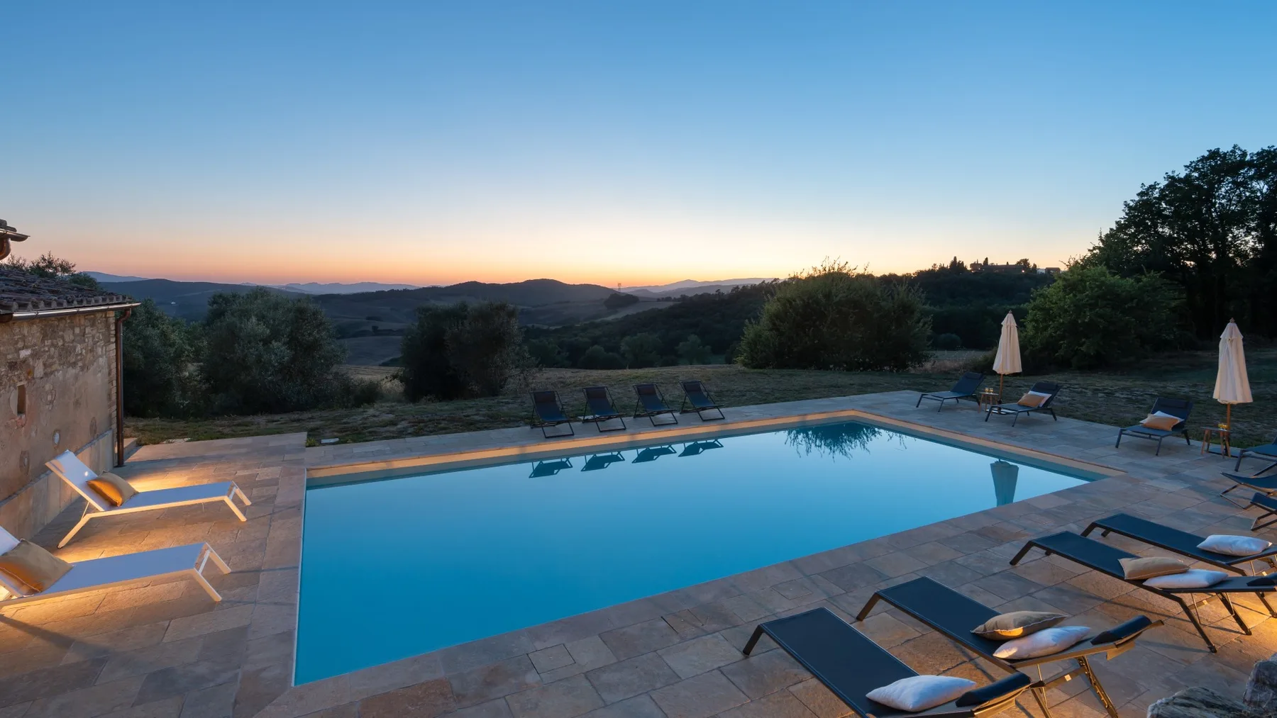 A swimming pool and lounge seating overlook a wooded landscape at sunset.