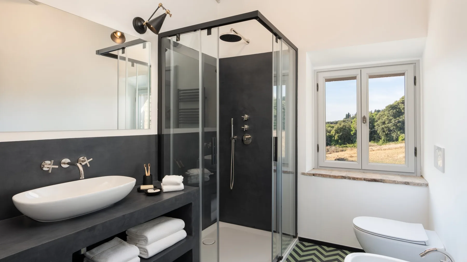 A bathroom with a glass shower, a vessel sink, and a window overlooking greenery.