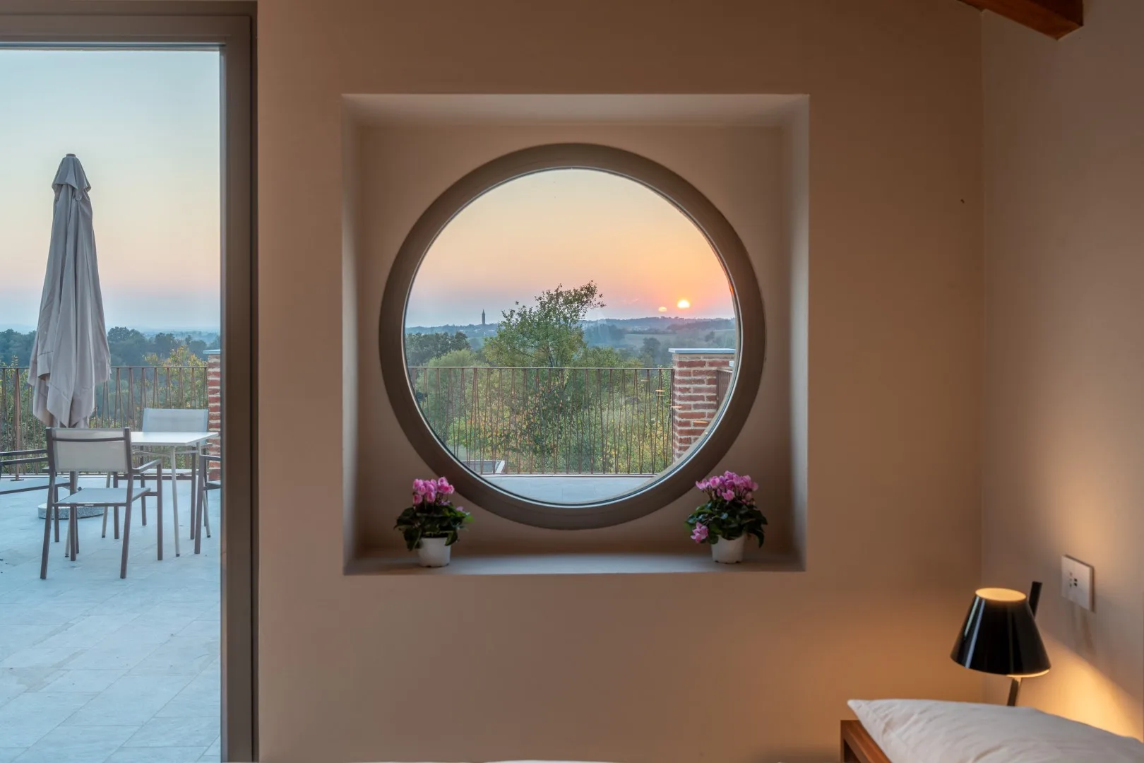 A bedroom with a round window overlooking a sunset and a terrace.