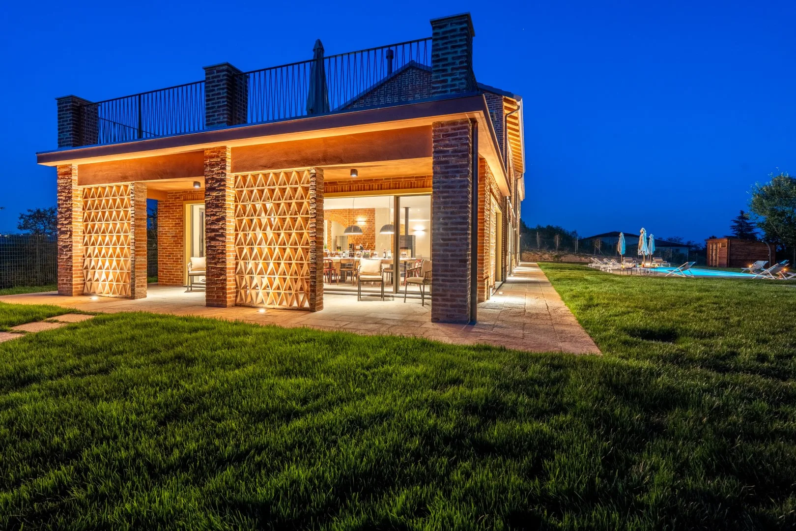 A modern house glows at dusk, with a rooftop terrace and lawn.