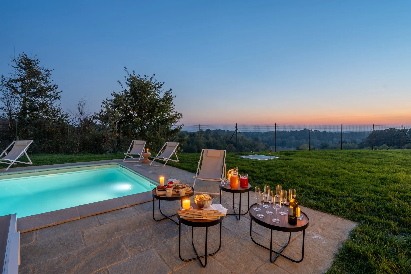 An outdoor poolside patio overlooks a grassy landscape at sunset.