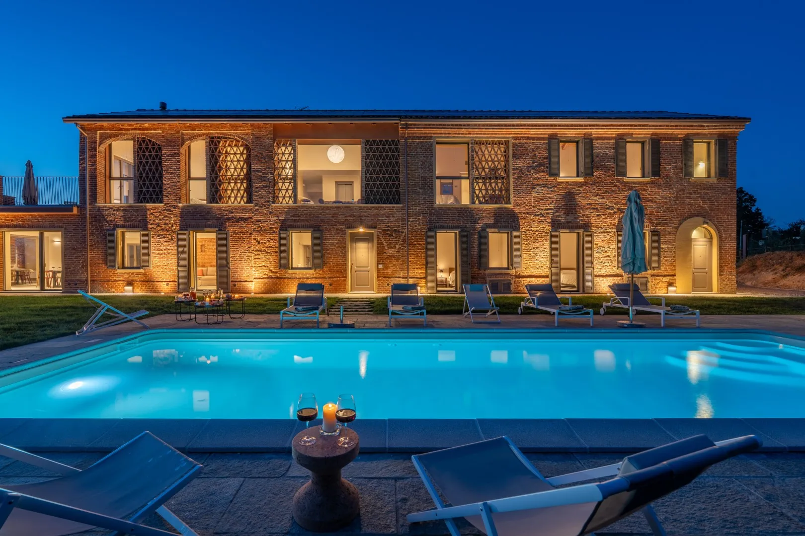 An illuminated brick building stands behind a swimming pool at dusk.