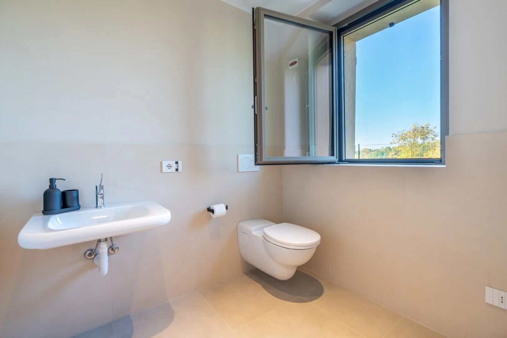 A bathroom with a wall-mounted sink, toilet, and open window.