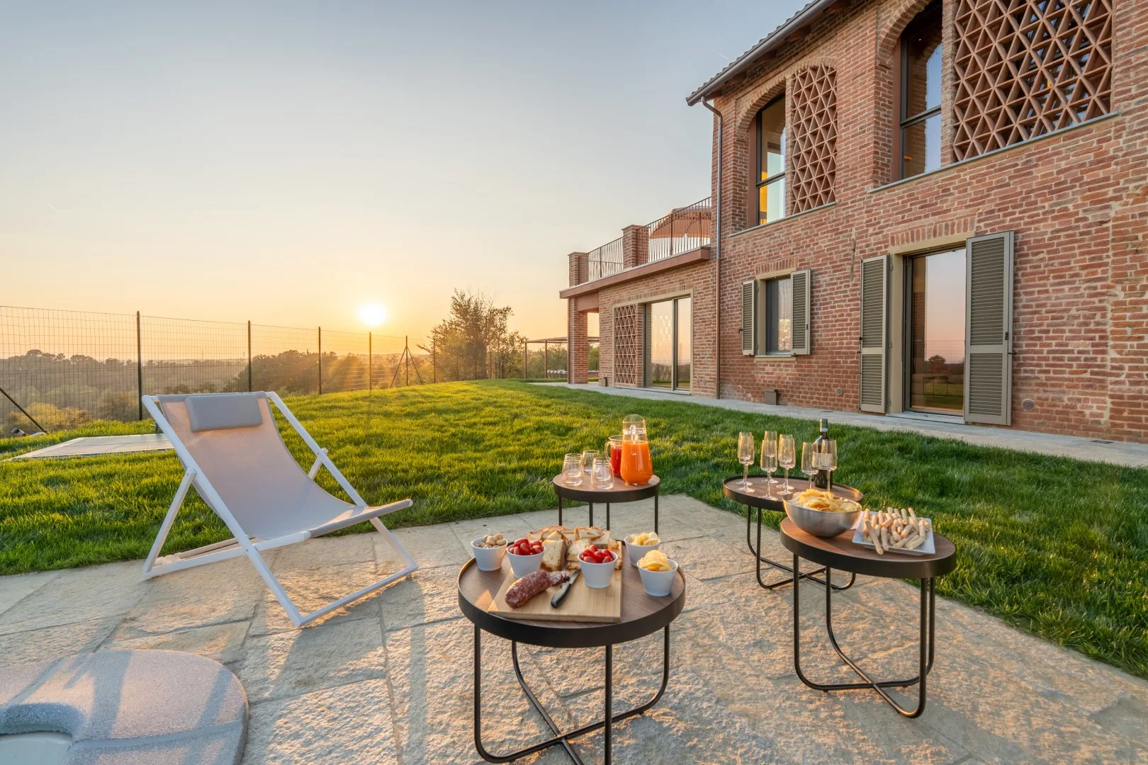 A brick house opens onto a grassy patio with outdoor seating and a sunset view.