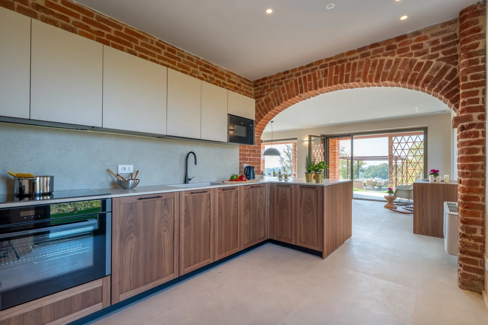 A modern kitchen with wood cabinetry opens through a brick arch to a bright dining area.