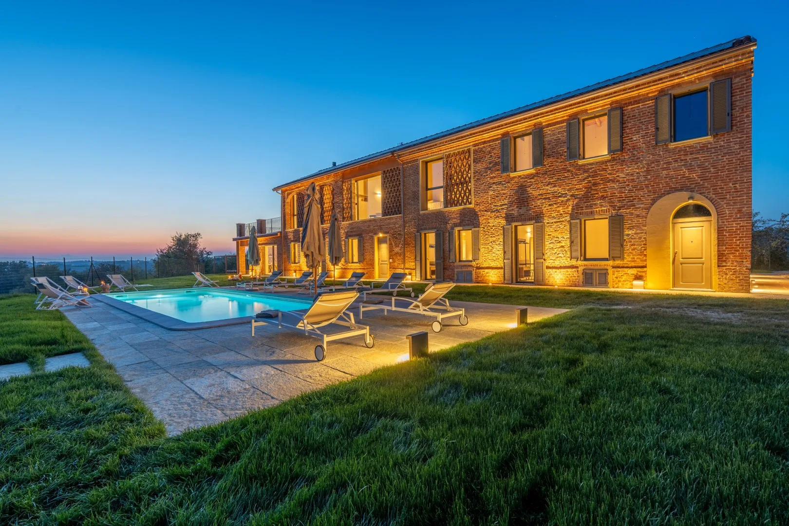 A brick building borders an outdoor pool and lawn at dusk.