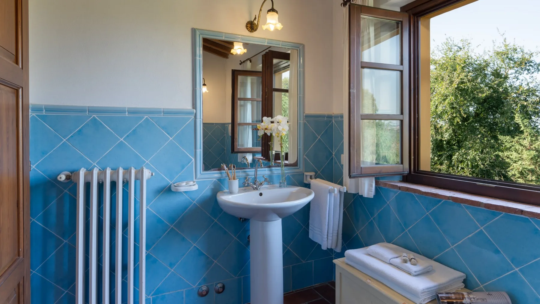 Un baño con lavabo de pedestal, paredes de azulejos azules y una ventana abierta.