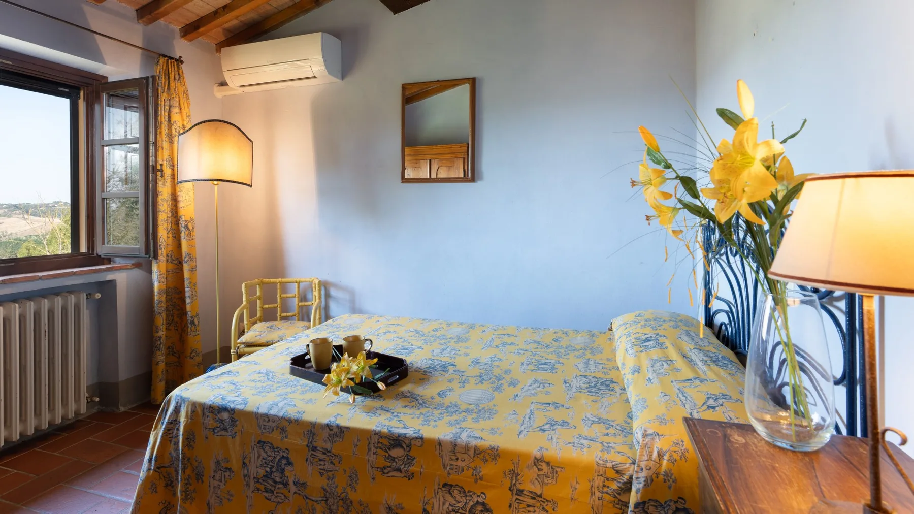 Un dormitorio con una cama cubierta con una colcha amarilla estampada y flores sobre una mesa.