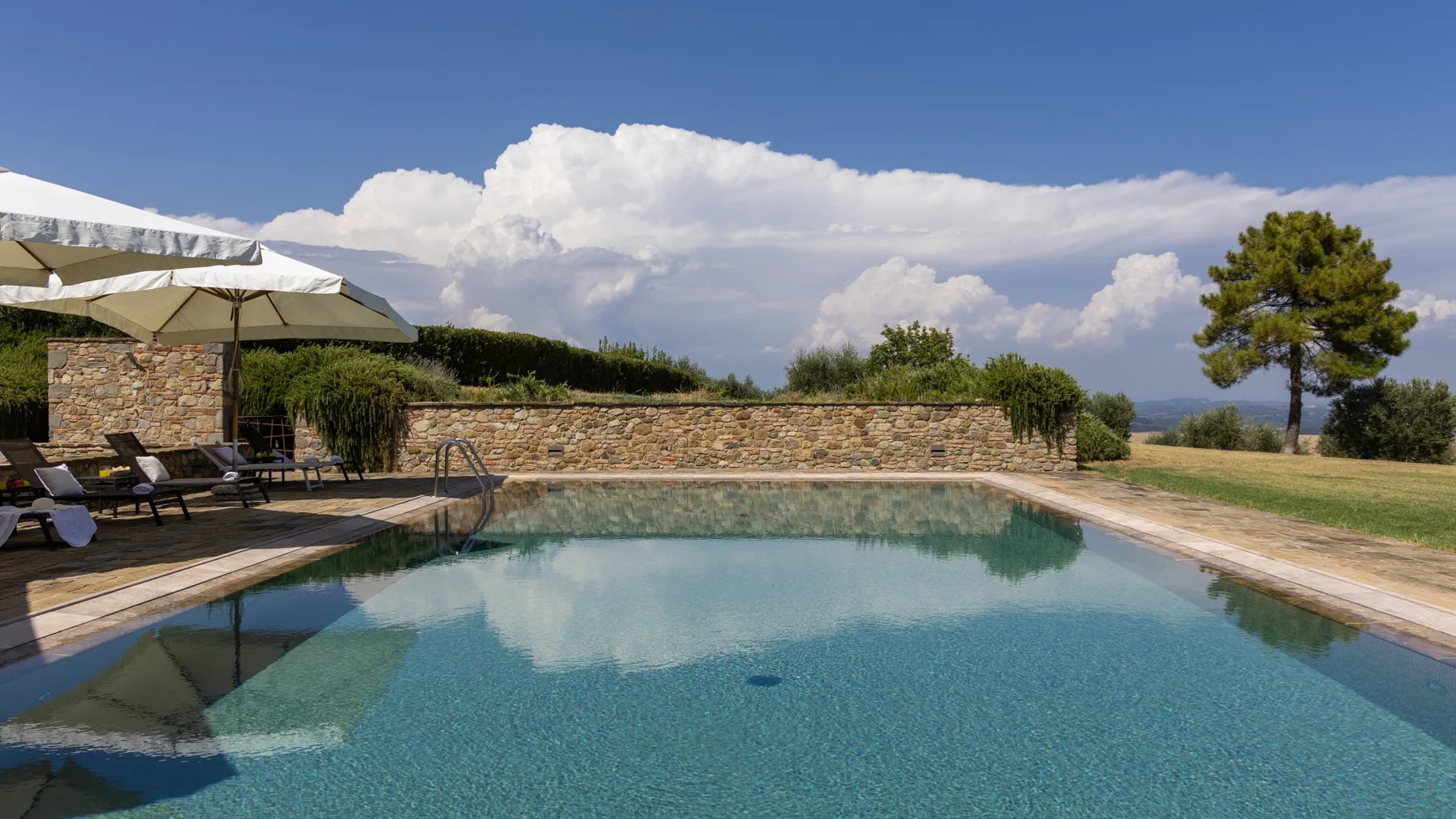 Una piscina al aire libre junto a un muro de piedra, con tumbonas y sombrillas cerca.