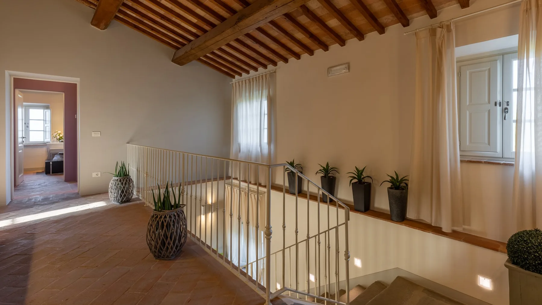 Un descansillo interior con barandilla metálica, vigas de madera vistas y ventanas.
