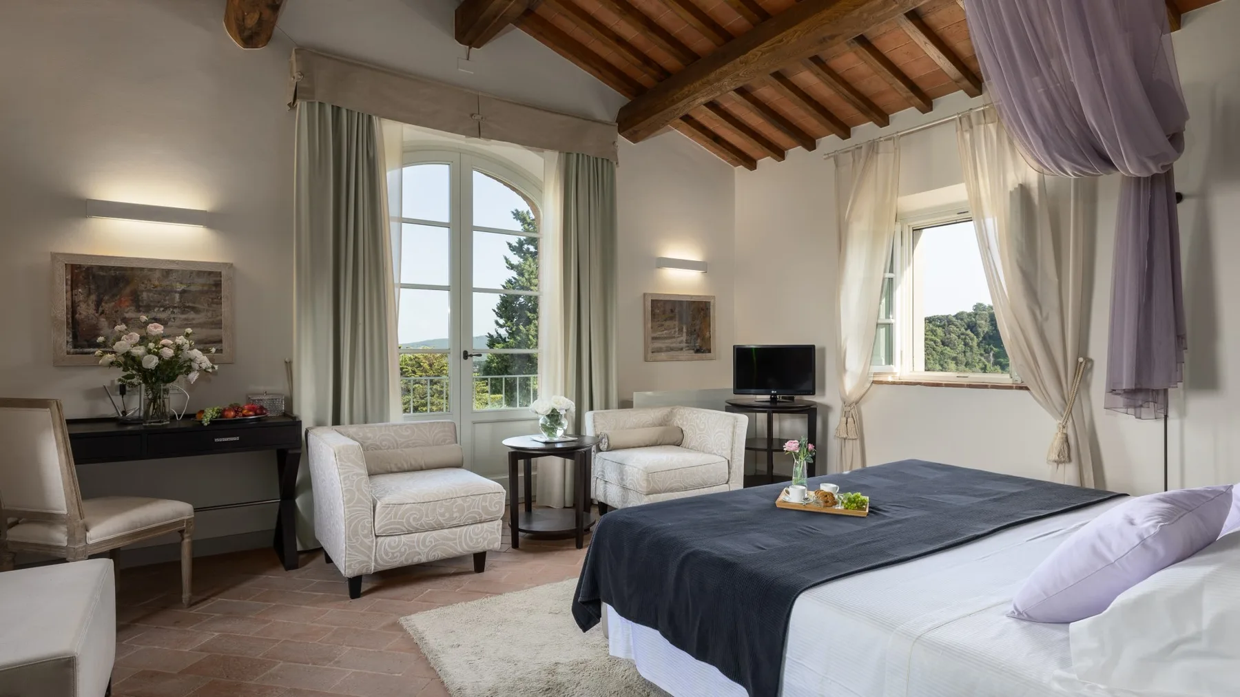Dormitorio luminoso con una cama grande, dos zonas de asiento y ventanas con vistas a la vegetación.