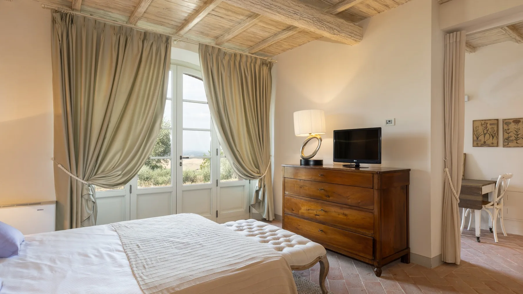 Un dormitorio con cama, cómoda y televisor junto a ventanas altas con cortinas.