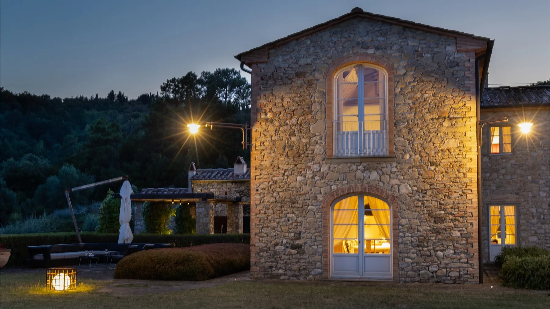 Una casa de piedra se alza junto a una terraza ajardinada, con luz cálida en las ventanas.