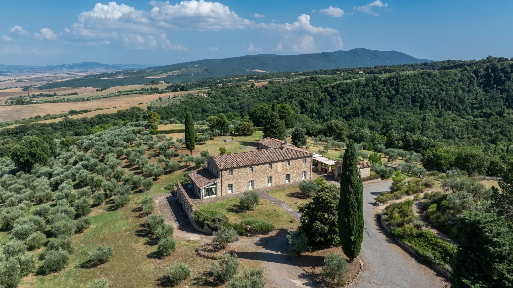 Una casa de piedra se encuentra entre olivares y colinas boscosas.