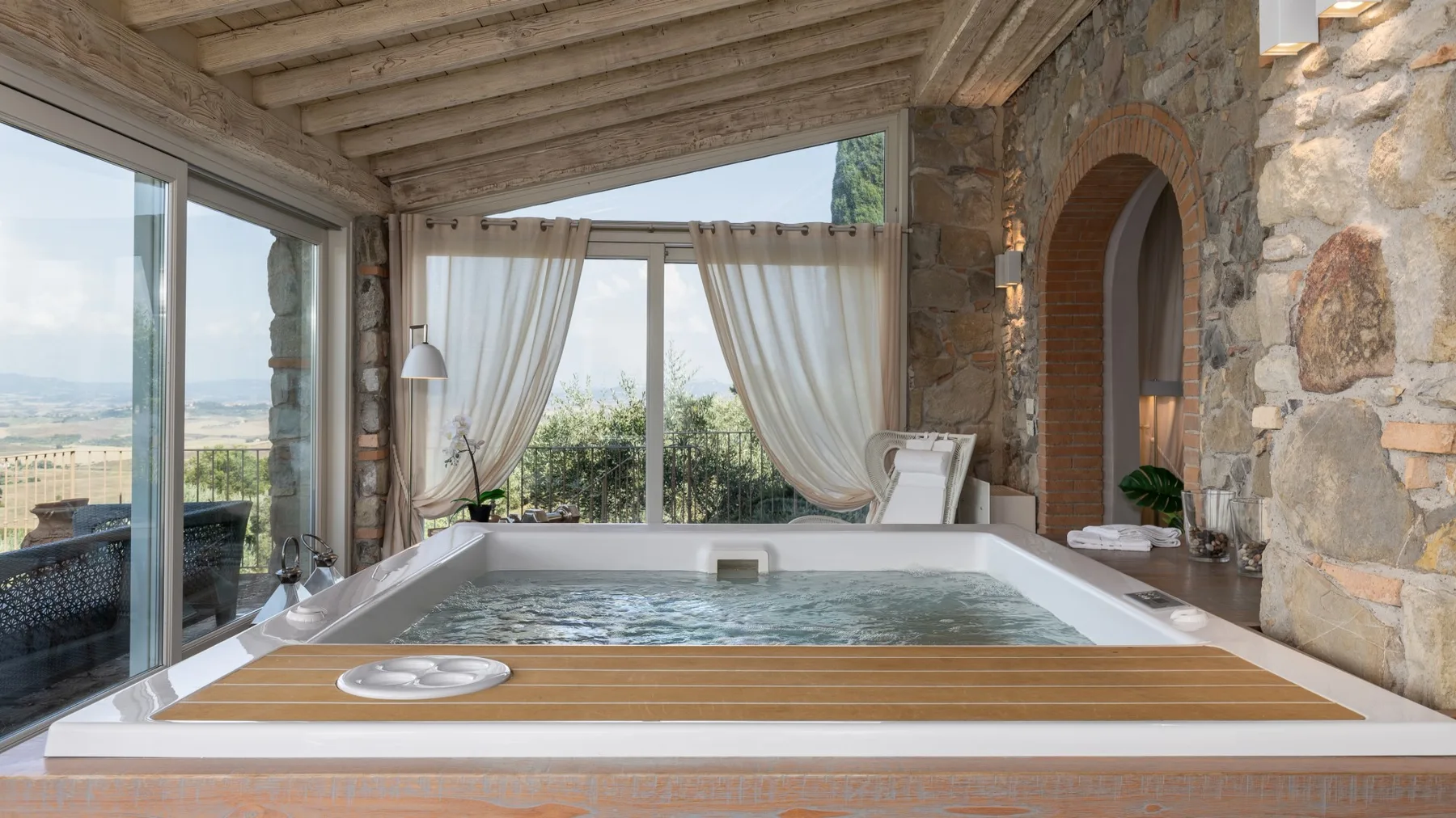 Un solárium con una bañera empotrada junto a grandes ventanales y paredes de piedra.