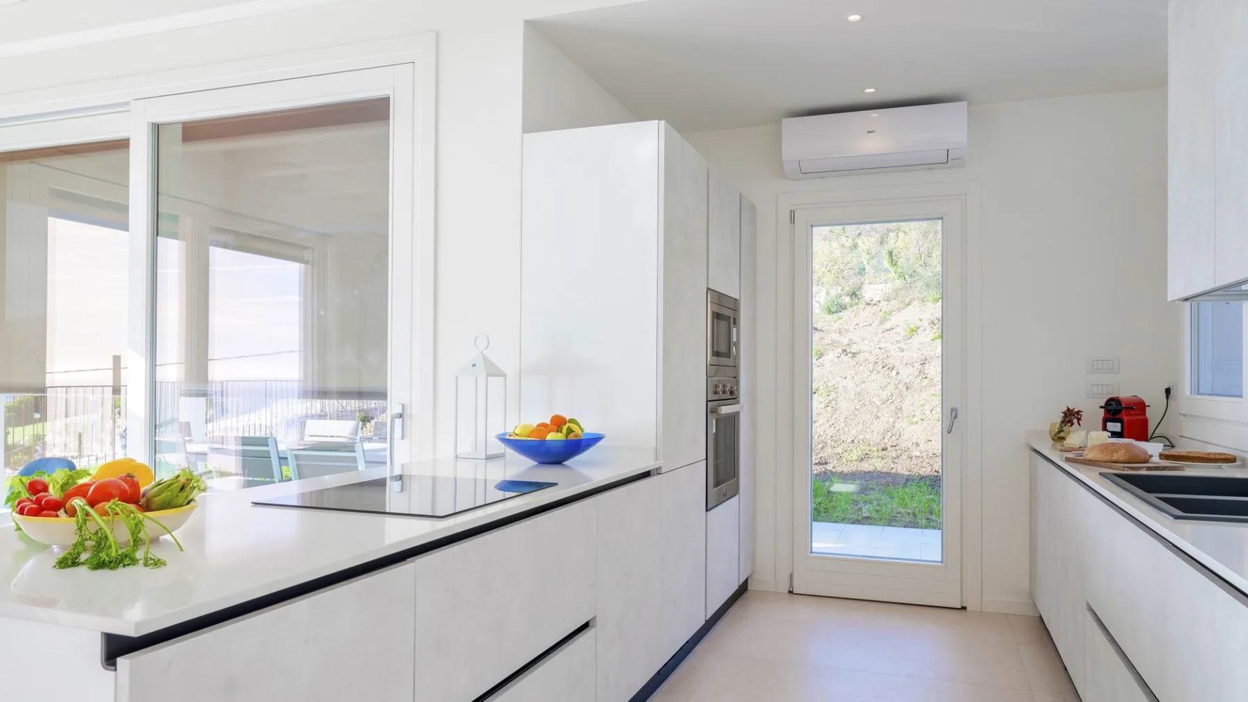 Una cocina blanca y luminosa con una isla central y una puerta de vidrio hacia la vegetación.