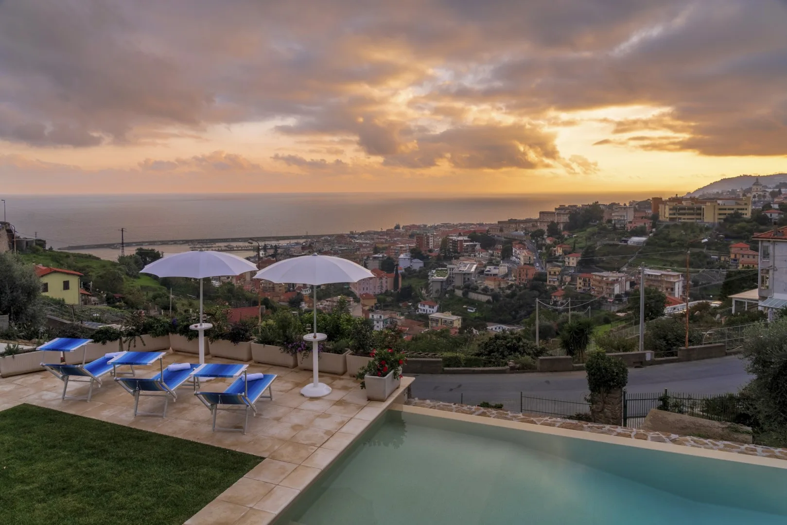Una terraza da a una piscina, un pueblo costero y el océano al atardecer.