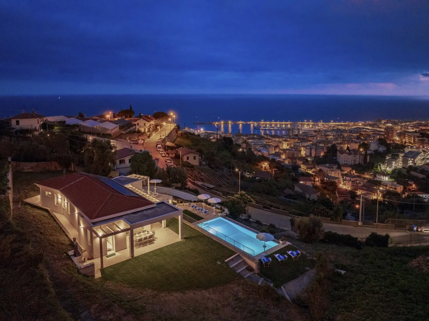 Una casa en la ladera y una piscina iluminada miran hacia un pueblo costero al anochecer.
