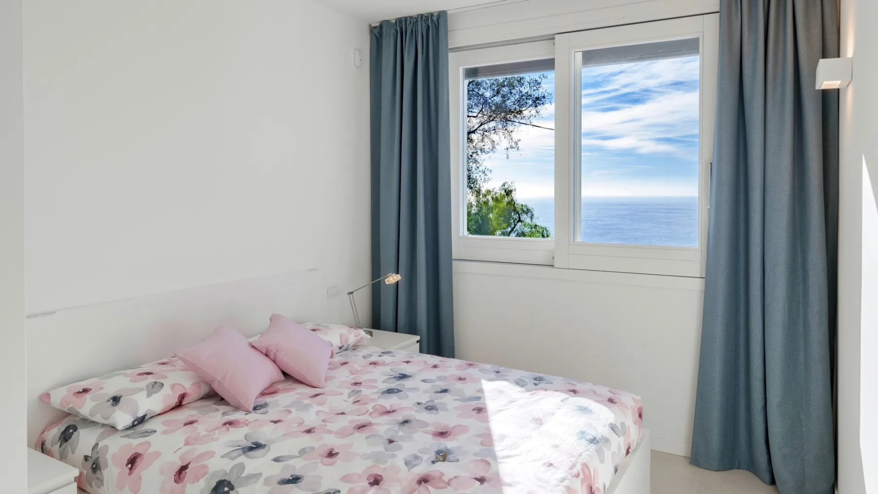 Un dormitorio con ropa de cama floral y cortinas azules junto a una ventana luminosa.