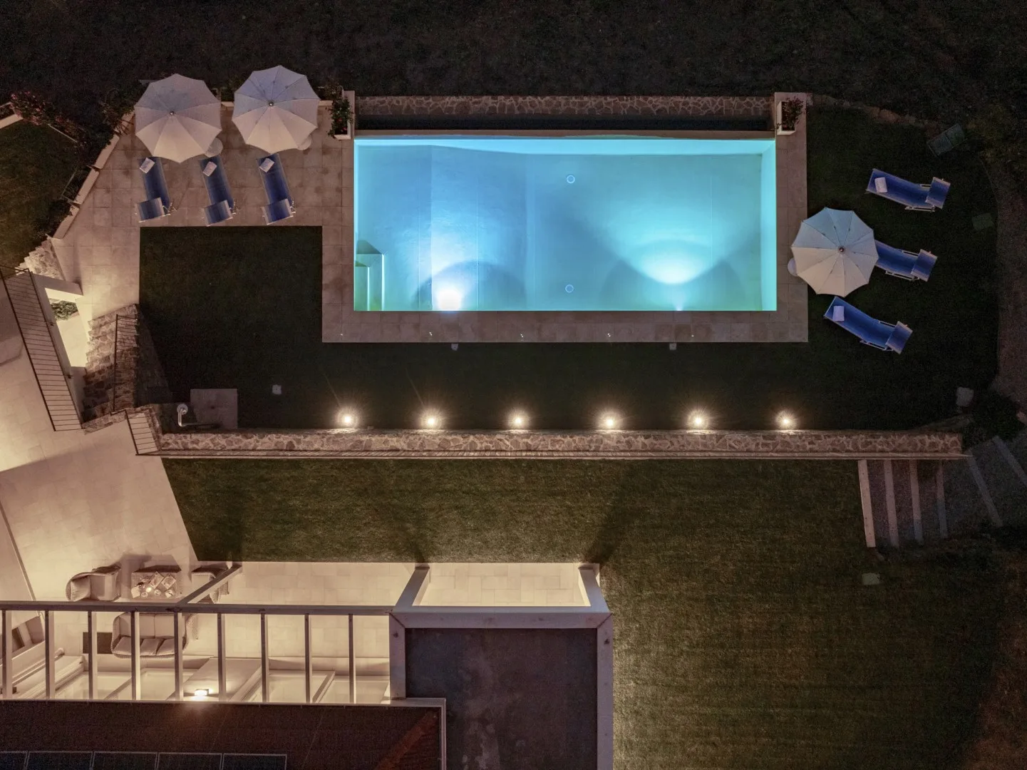 Una piscina iluminada está junto a un césped y asientos al aire libre por la noche.