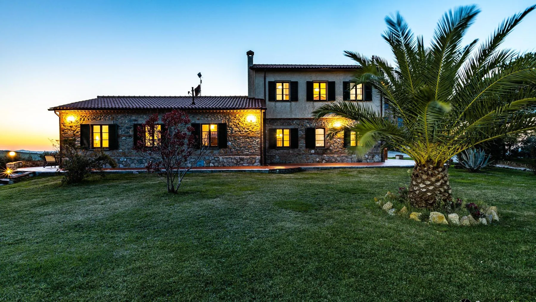 A large house with warmly lit windows stands behind a lawn and palm tree at dusk.