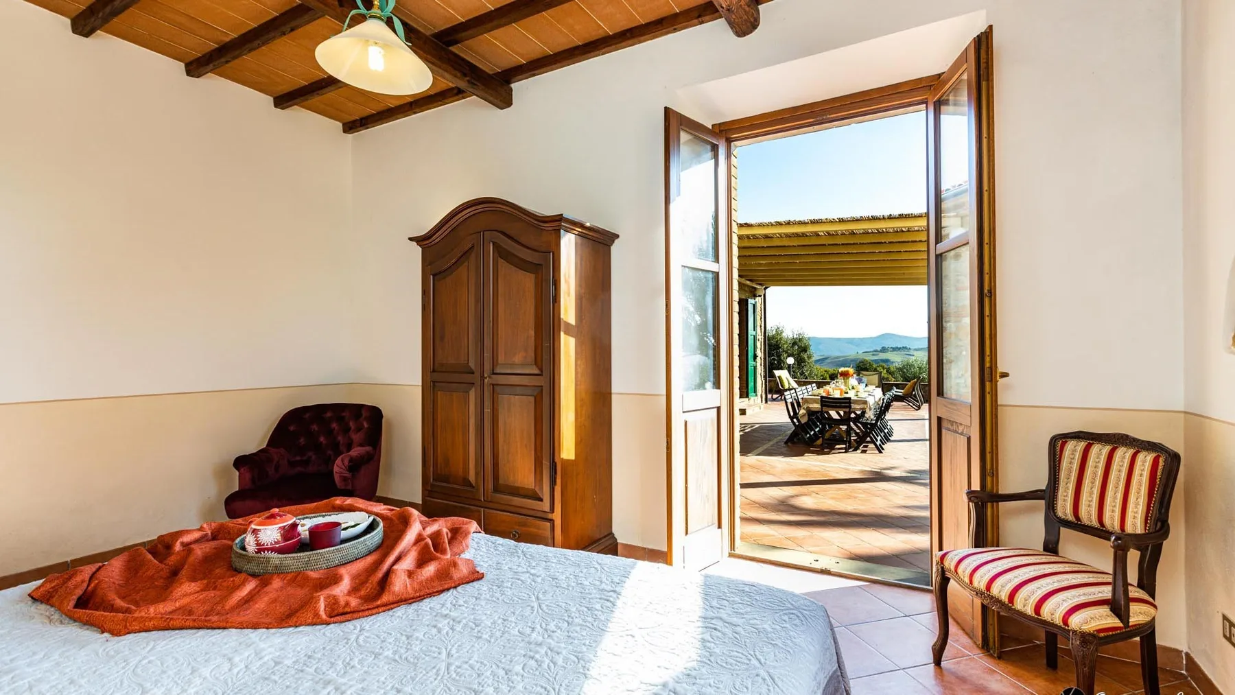 A bedroom opens through double doors onto a sunlit terrace, with seating and a wardrobe nearby.