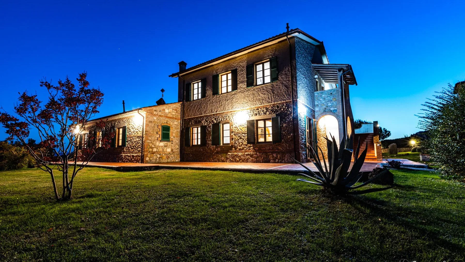 A stone house is illuminated at dusk beside a grassy lawn.