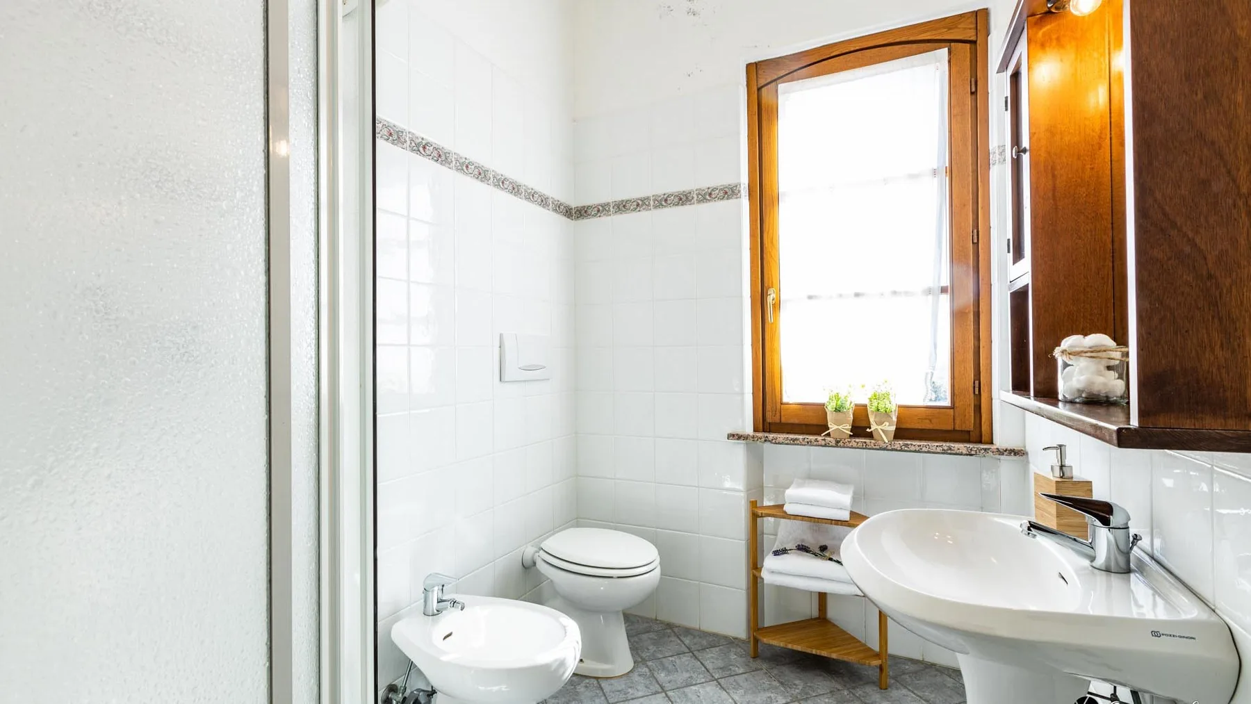 A bright tiled bathroom with a sink, toilet, and window.