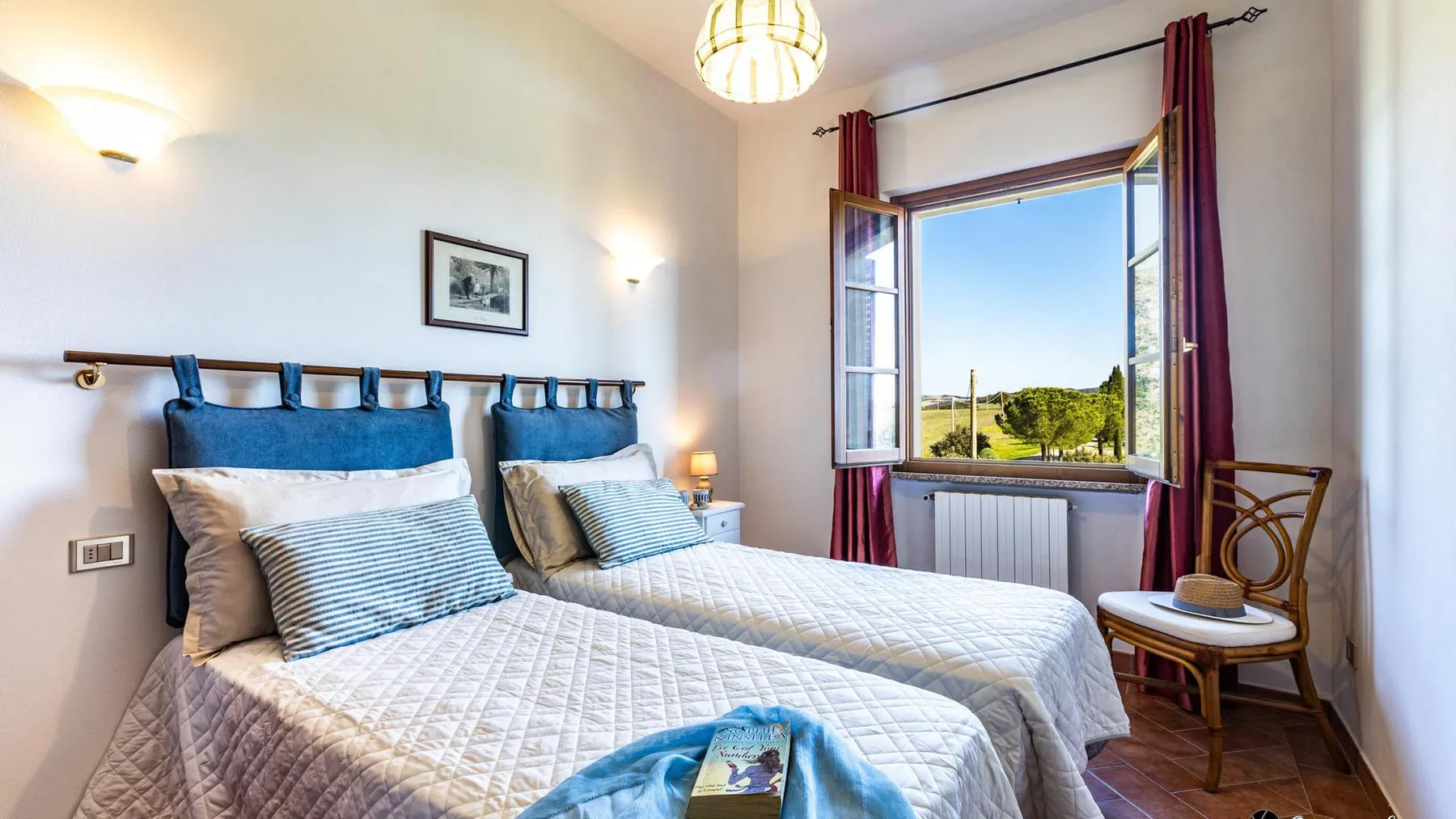 A bedroom with two beds beside an open window overlooking greenery.