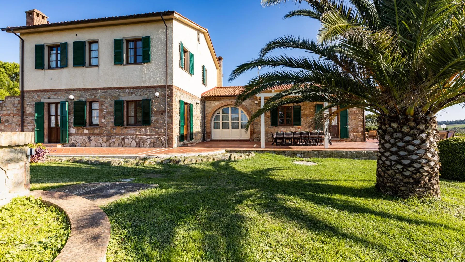 A two-story house with a tiled roof stands beside a palm tree and lawn.