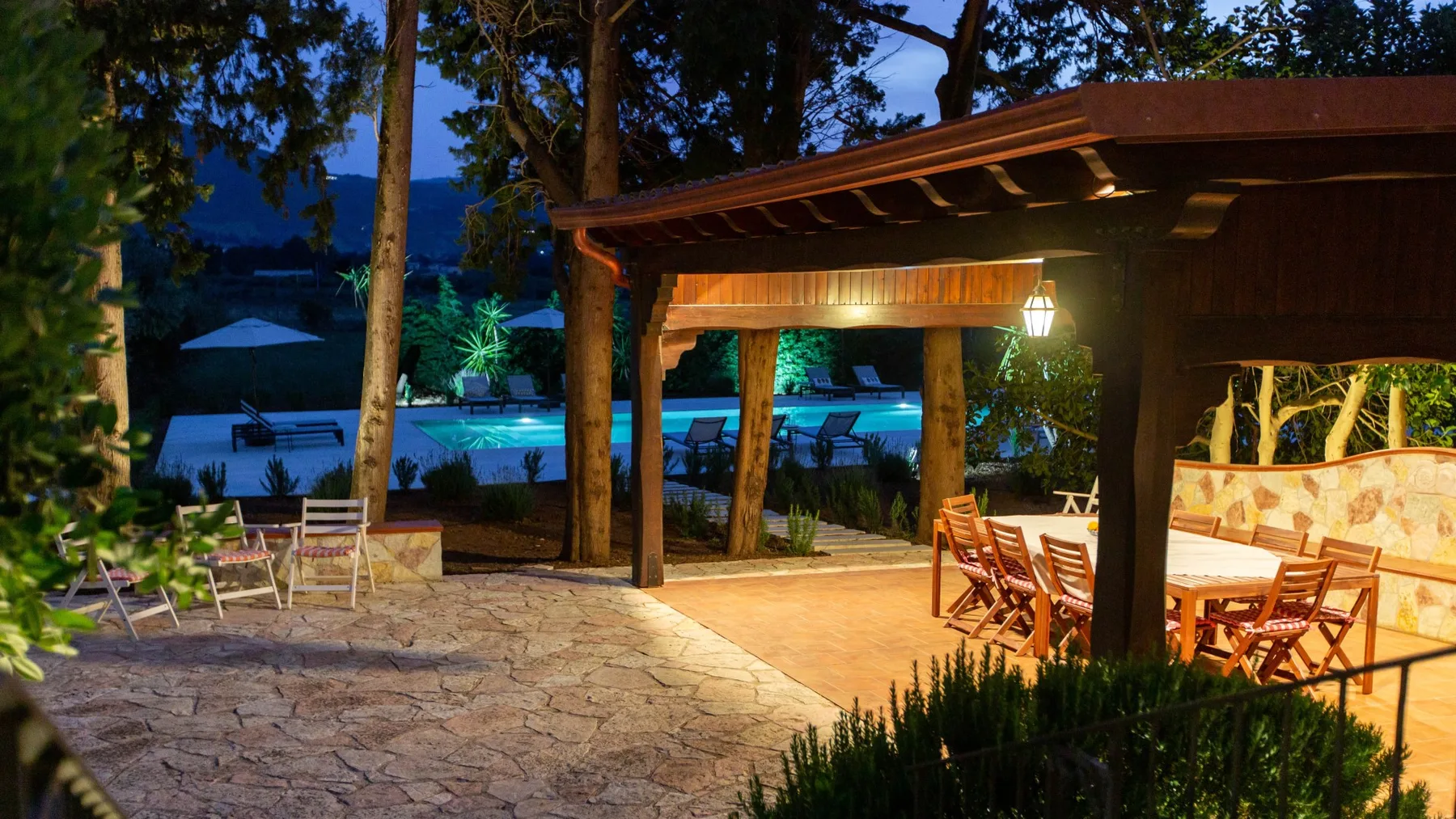 Una zona de comedor al aire libre bajo una pérgola de madera da a una piscina iluminada.