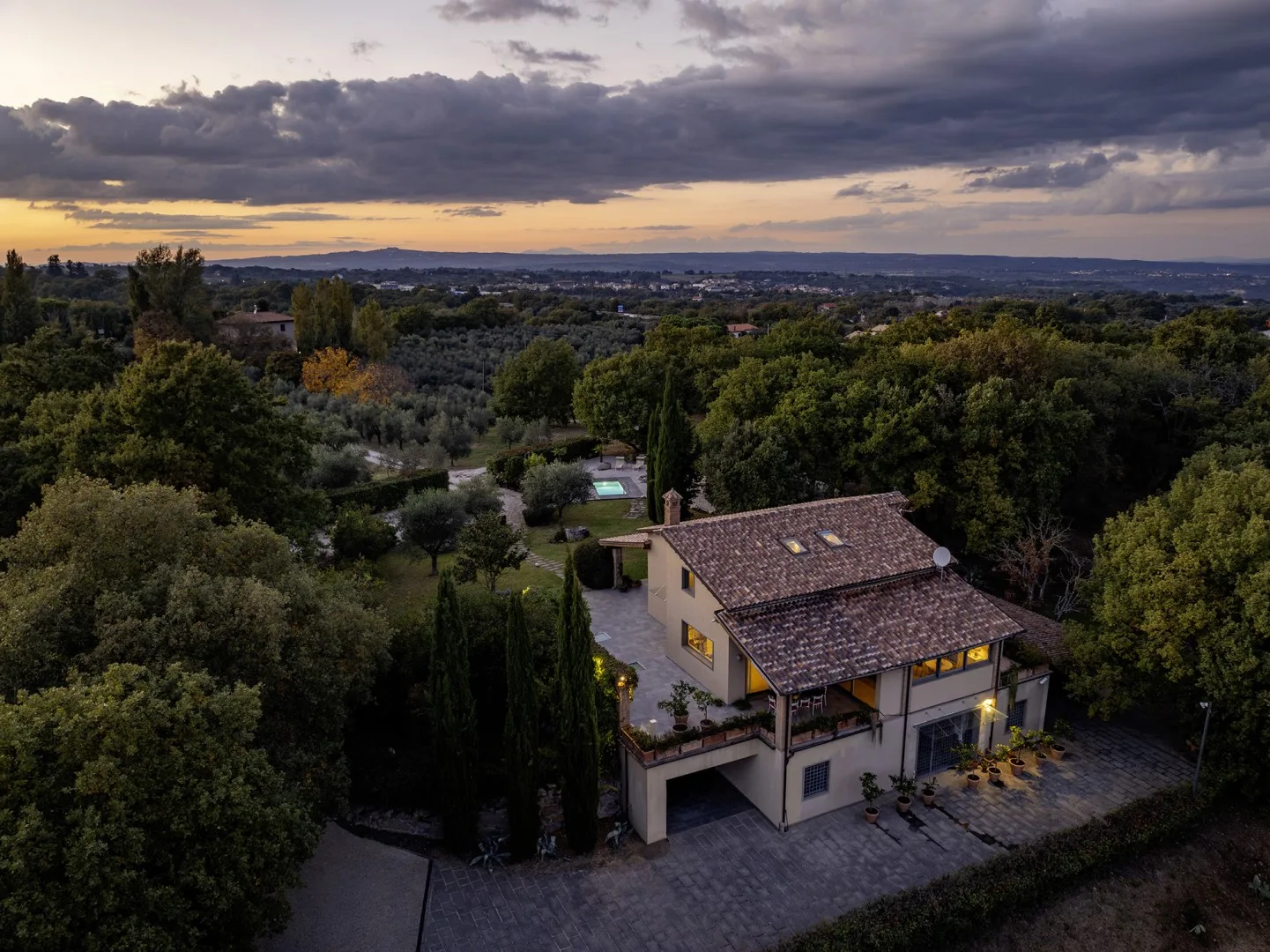 A countryside house sits among trees beneath a cloudy sunset sky.