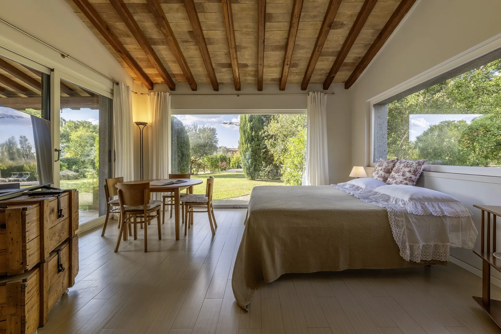 A bright bedroom with a bed, dining table, and large windows overlooking greenery.