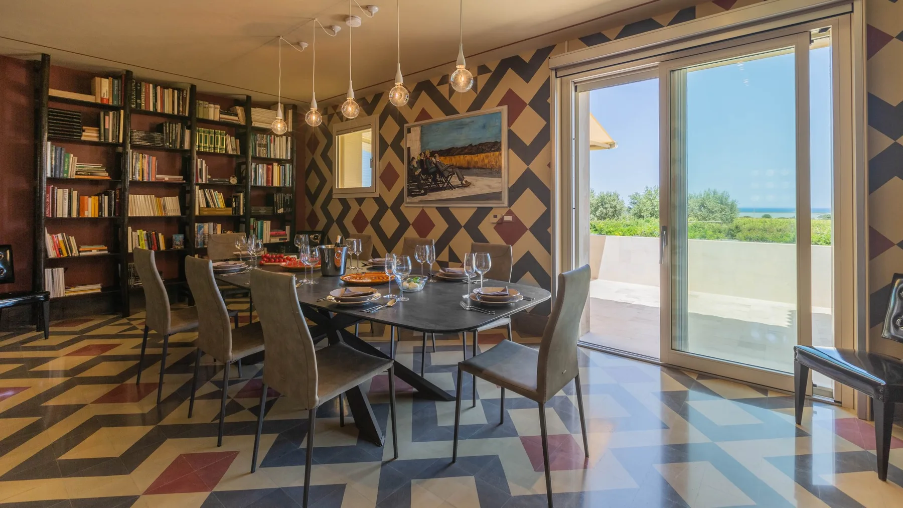 A dining area with a set table, bookshelves, and large windows overlooking greenery.