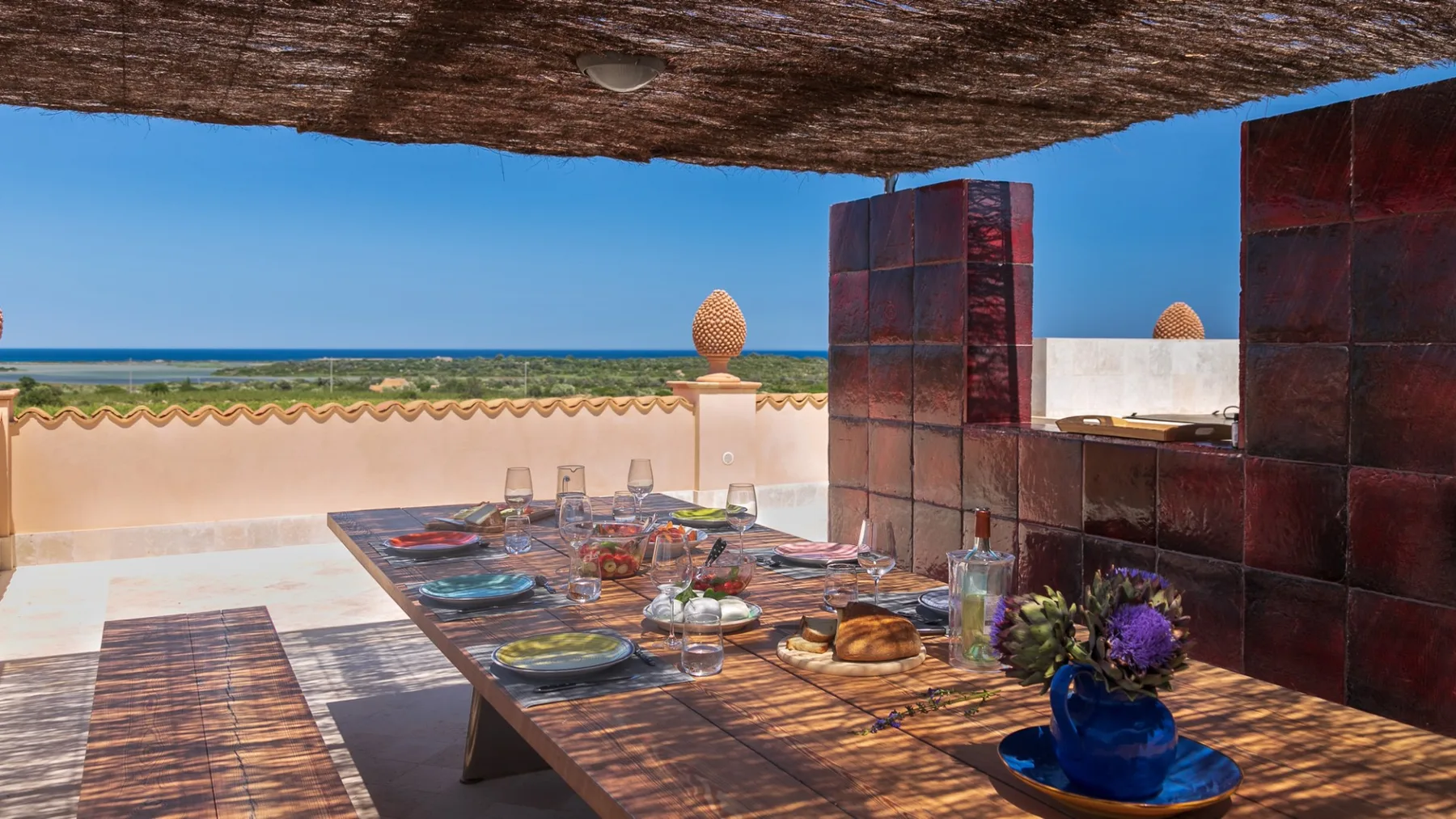 An outdoor dining table is set beneath a shaded canopy, with the sea visible beyond.