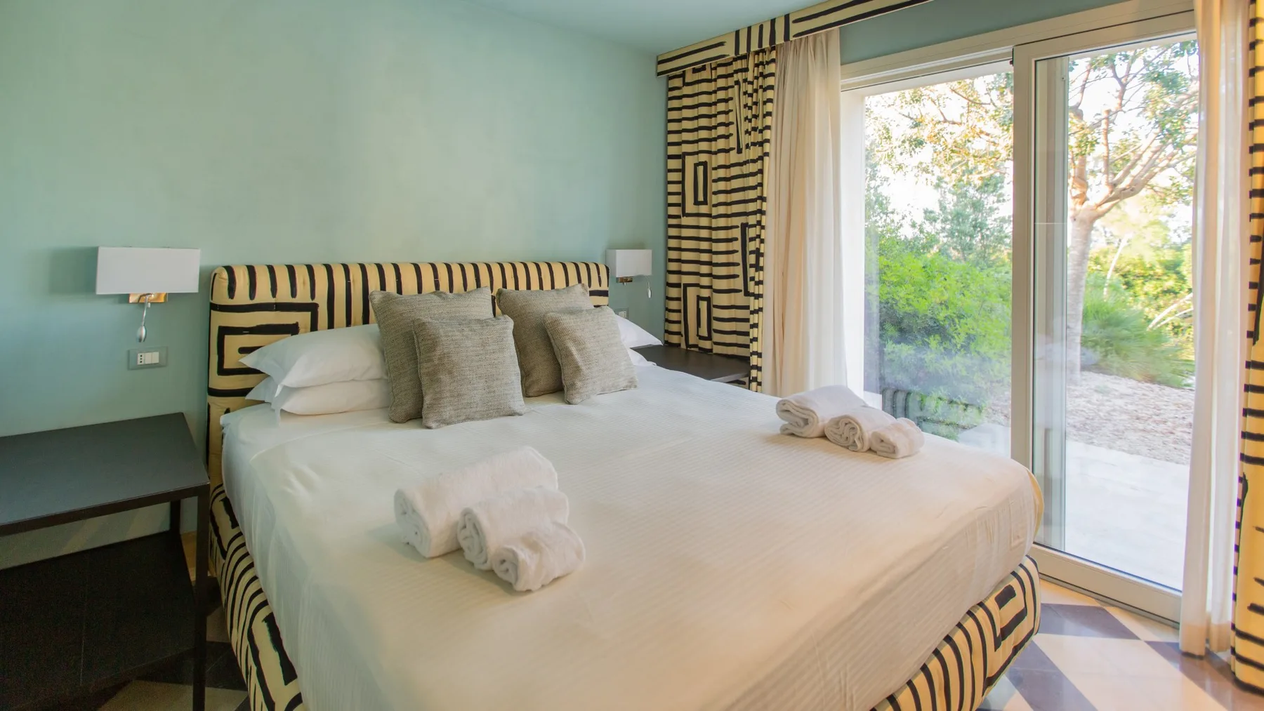 A bedroom with a large bed beside glass doors opening onto greenery.
