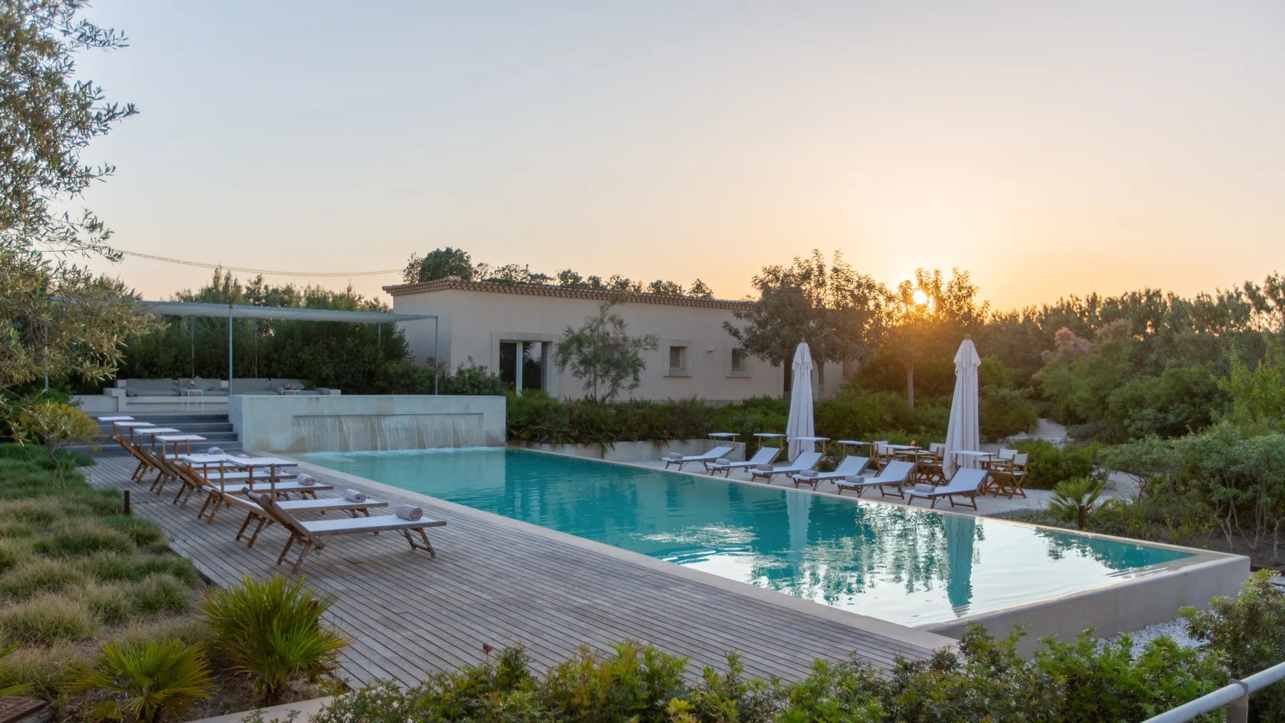 An outdoor swimming pool with lounge seating sits beside a low building at sunset.